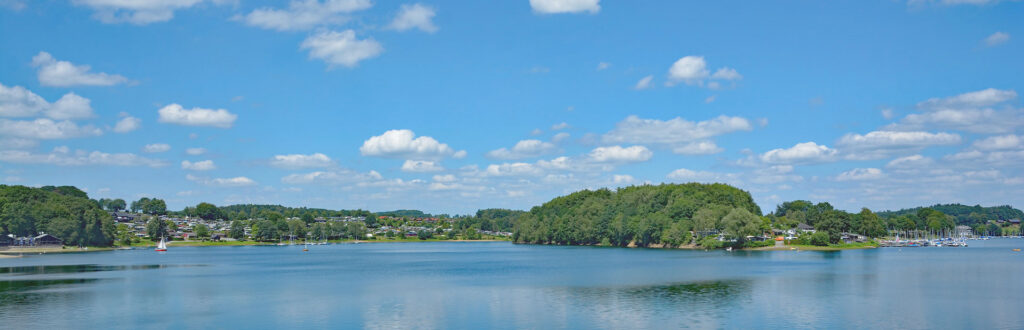 Panorama eines Sees mit blauem Himmel, weißen Wolken und bewaldeten Ufern.