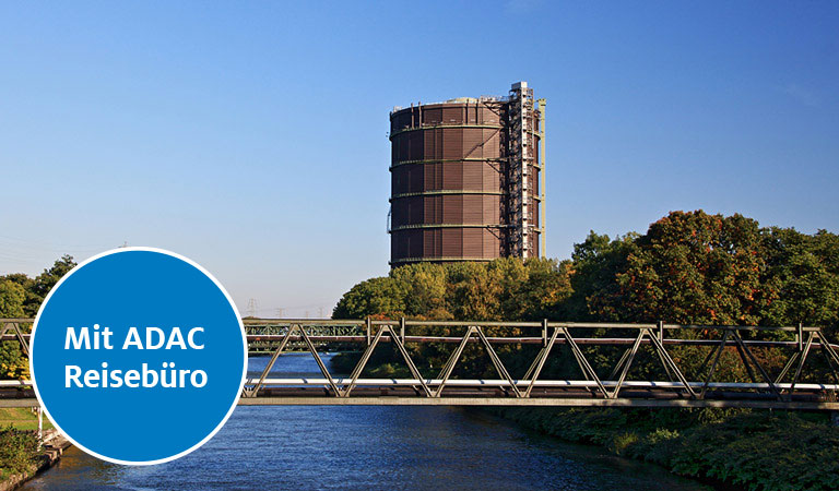 Gasometer in Oberhausen bei klarem Himmel, mit Brücke im Vordergrund und Herbstbäumen.