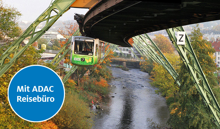Ein grüner Schwebebahnwagon fährt über einen herbstlich gefärbten Fluss.