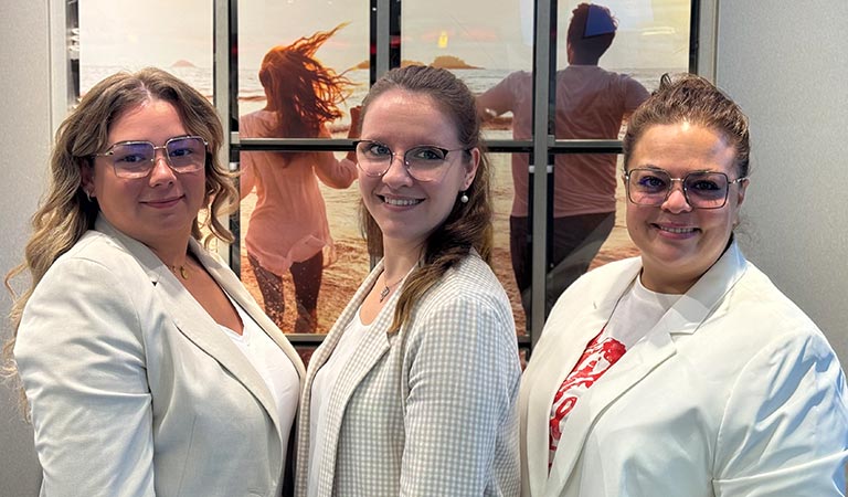 Drei lächelnde Frauen in weißer Kleidung stehen vor einem Strandbild.