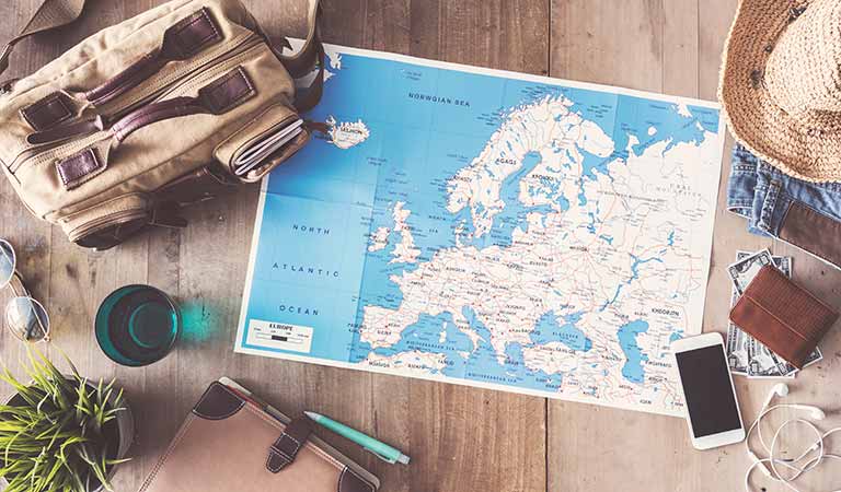 Eine Europakarte liegt auf einem Holztisch neben Reisetasche, Handy und Sonnenhut