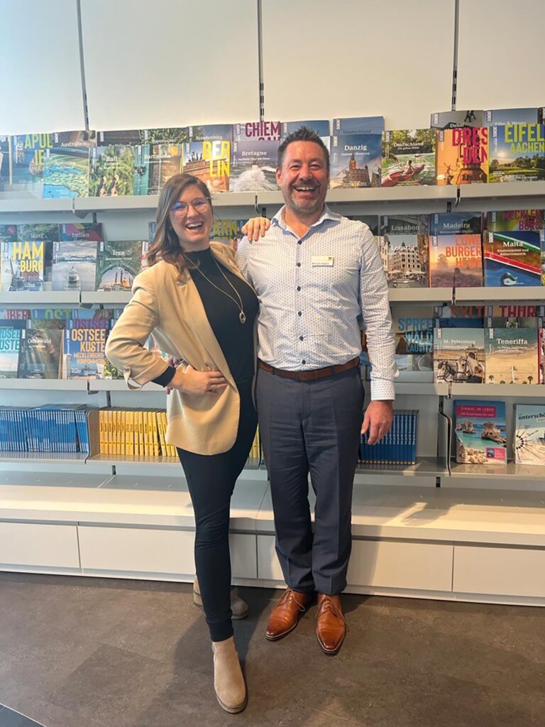 Eine lächelnde Frau und ein lachender Mann stehen vor bunten Reiseführern im Regal