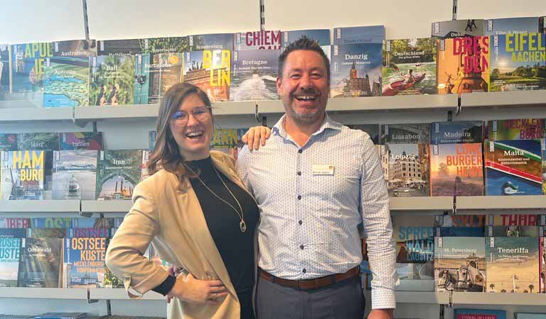 Ein lachender Mann im blauen Hemd steht neben einer Frau vor bunten Reiseführern