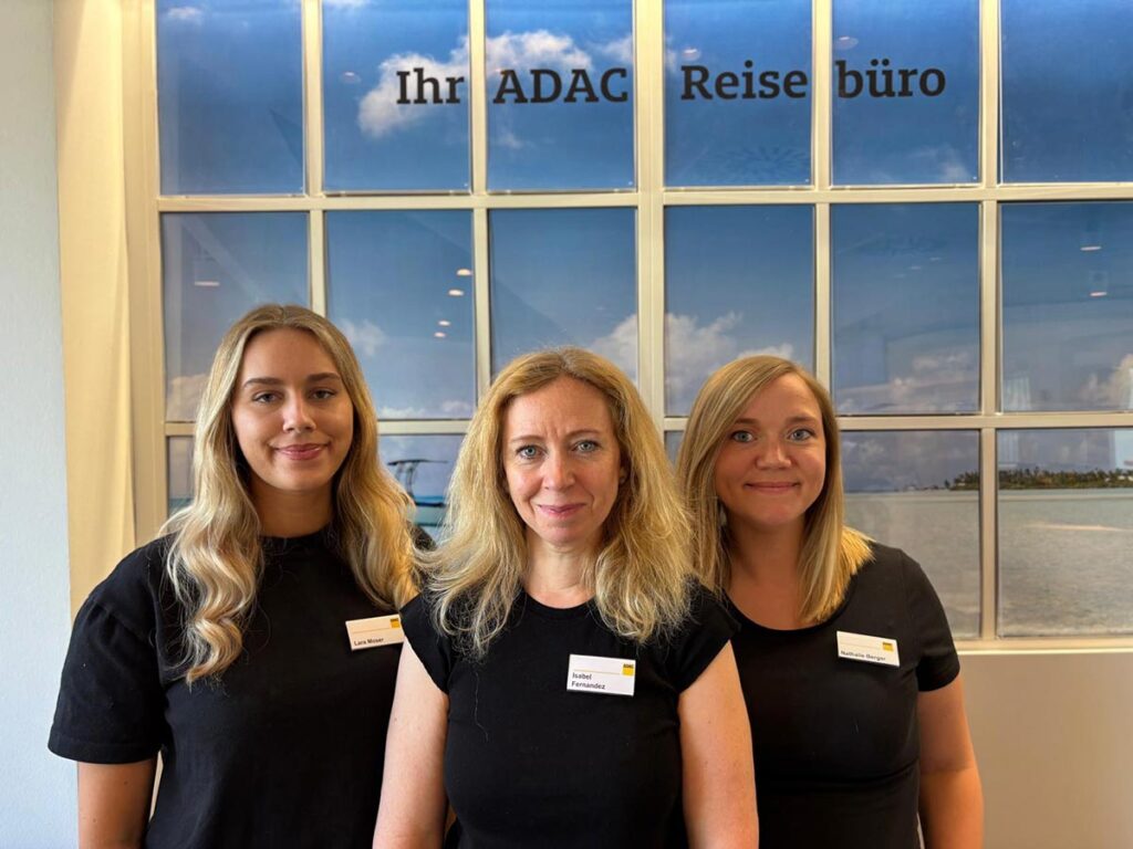 Drei Frauen mit Namensschildern lächeln im ADAC Reisebüro vor einem Fenster