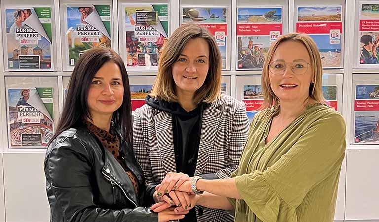 Drei Frauen mit freundlichen Gesichtern stehen nebeneinander und halten sich an den Händen