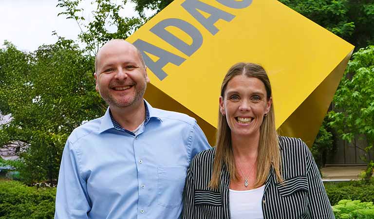 Ein lächelnder Mann und eine lächelnde Frau stehen vor einem gelben ADAC-Würfel