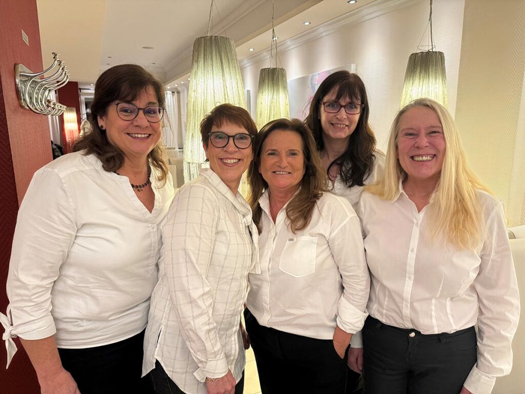 Fünf Frauen mittleren Alters lächeln freundlich in weißer Kleidung unter Leuchten