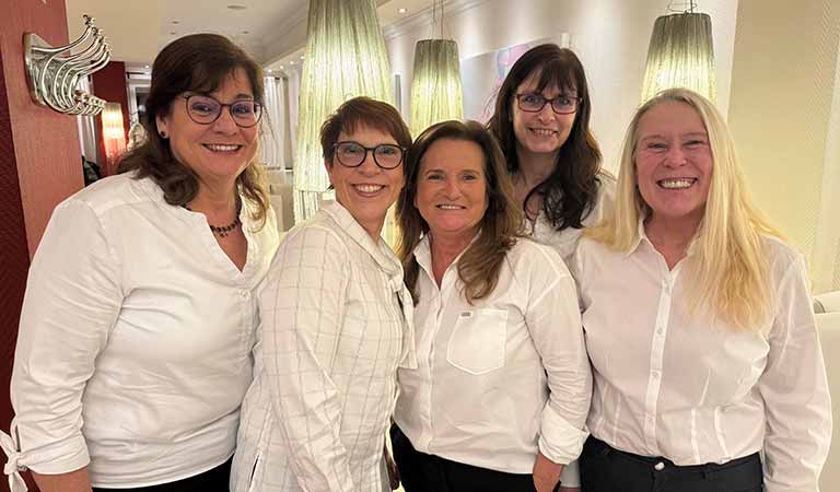 Fünf lächelnde Frauen mit Brillen und weißen Hemden stehen in einem hellen Raum