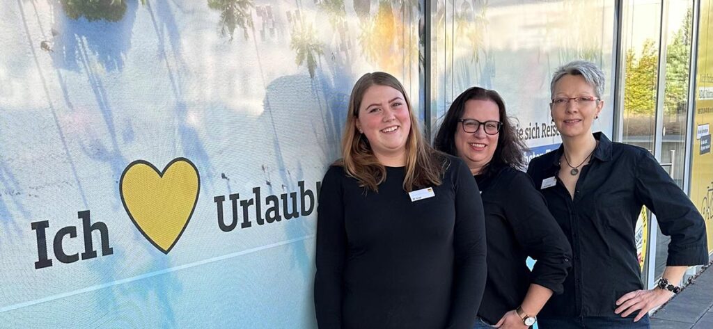 Drei Frauen stehen lächelnd nebeneinander vor einer Wand mit Urlaubswerbung