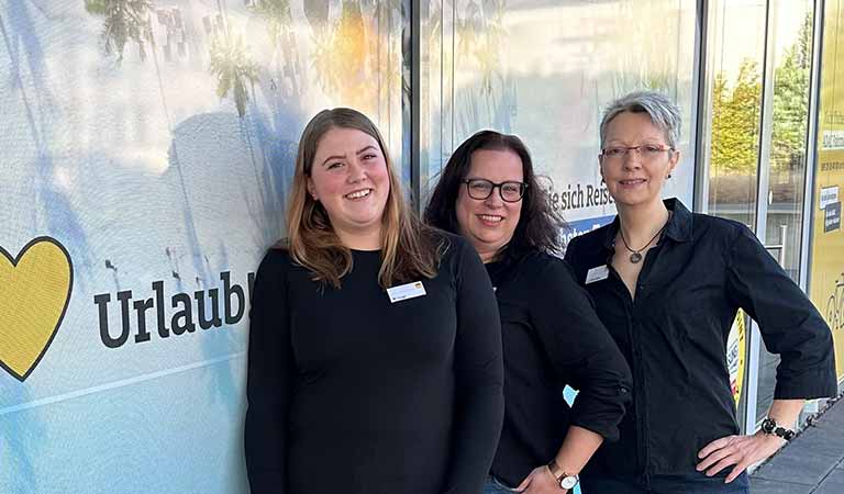 Drei Frauen mit Namensschildern lächeln freundlich vor einer sonnigen Glasfassade