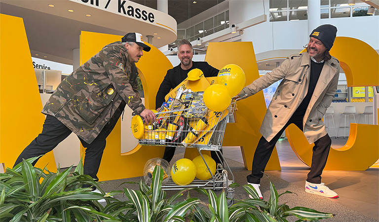 Drei Männer lachen fröhlich und ziehen an einem Einkaufswagen voller gelber Luftballons