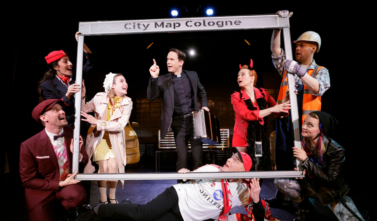 Eine Gruppe von acht bunt gekleideten Schauspielern posiert lebhaft um einen Rahmen mit der Aufschrift City Map Cologne, ihre Mimik reicht von neugierig bis amüsiert