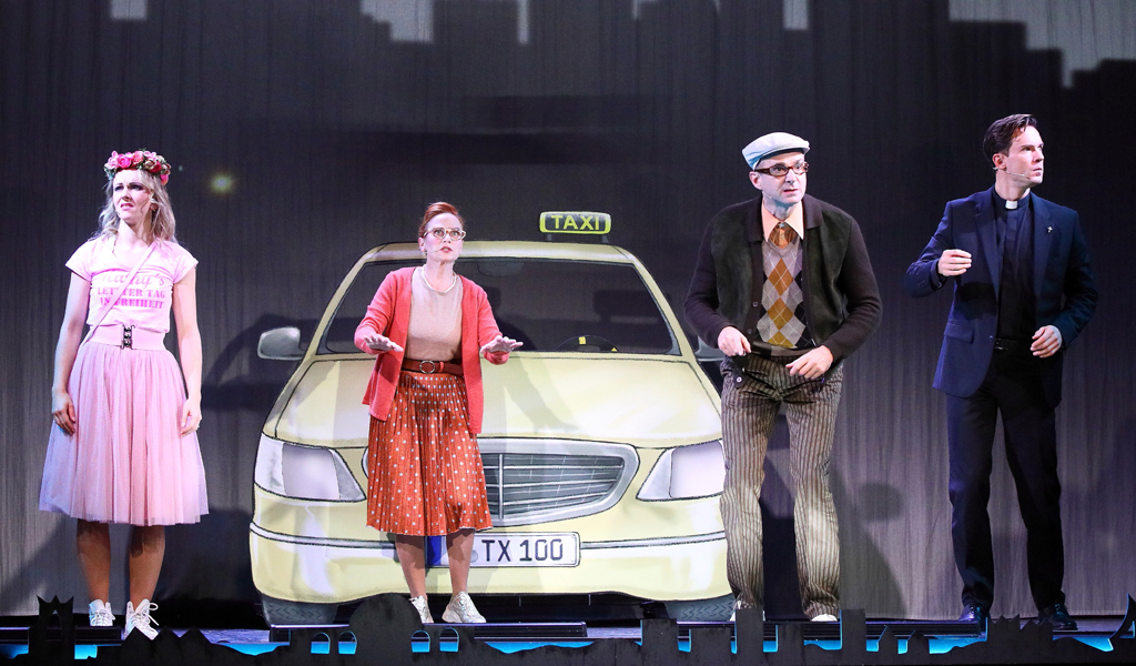 Vier Personen stehen vor einem gemalten Taxi, wirken überrascht und tragen bunte Kostüme