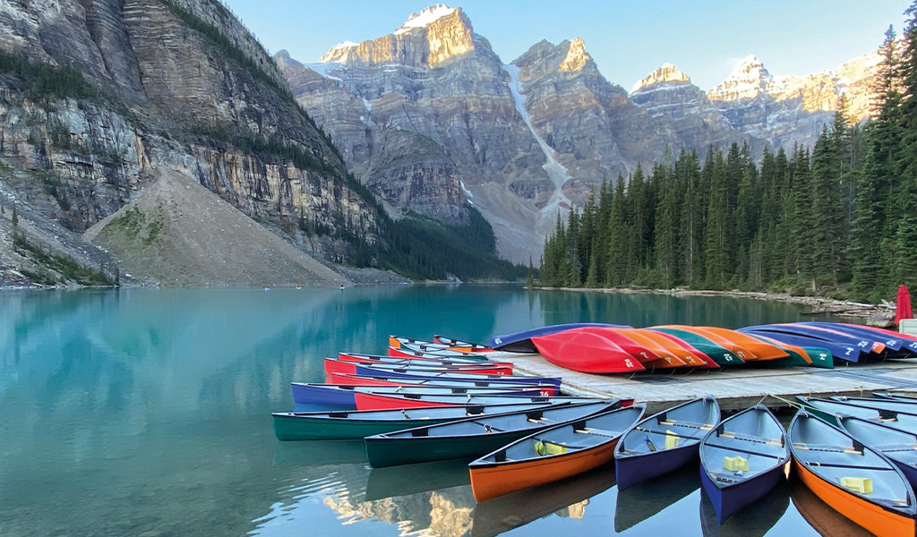 Mehrfarbige Kanus liegen auf einem Holzsteg an einem türkisfarbenen See vor Bergen