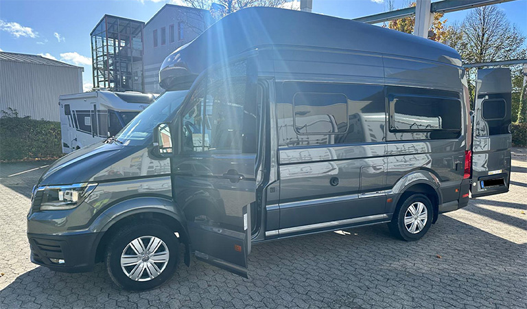 Ein dunkelgrauer Hochdach-Van mit offenen Türen steht auf einem sonnigen Parkplatz