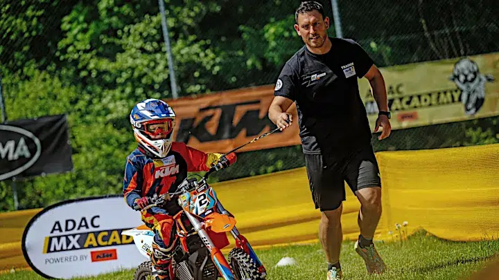 Ein Erwachsener mit schwarzem Shirt führt ein Kind in bunter MX Ausrüstung auf einem kleinen Motorrad