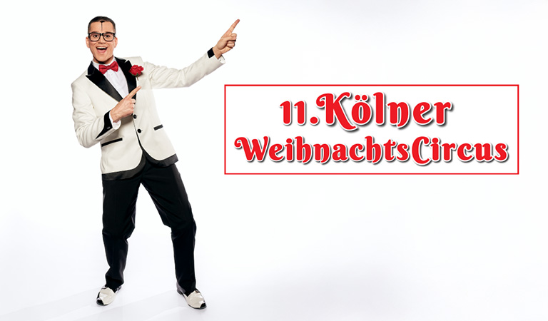 Ein lächelnder Mann im weißen Smoking mit roter Fliege zeigt auf festlichen Schriftzug