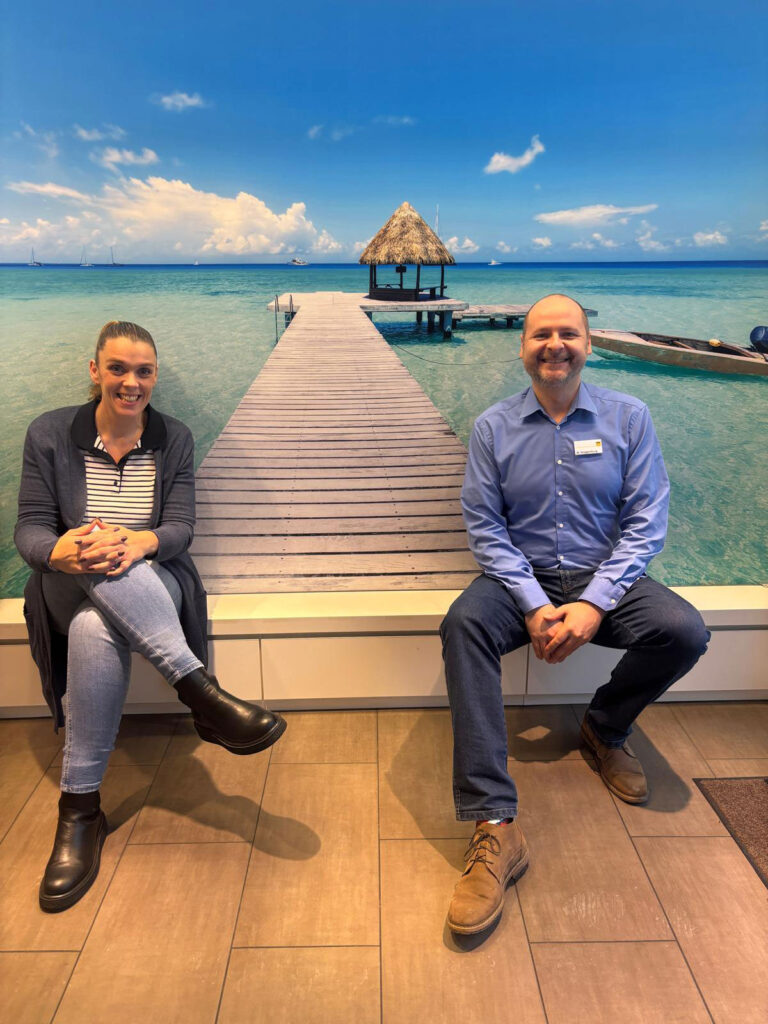 Zwei lächelnde Personen sitzen vor einem Wandbild mit Steg im türkisfarbenen Meer