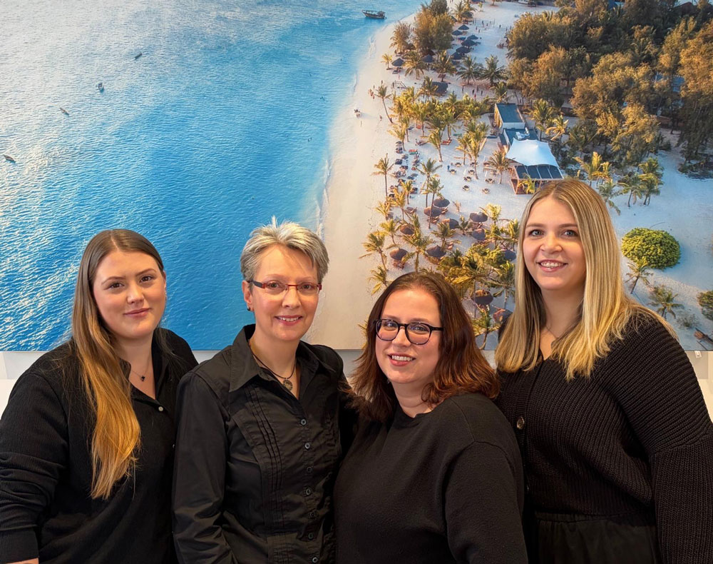 Vier Frauen in dunkler Kleidung lächeln vor einem Strandfoto mit Palmen und Meer