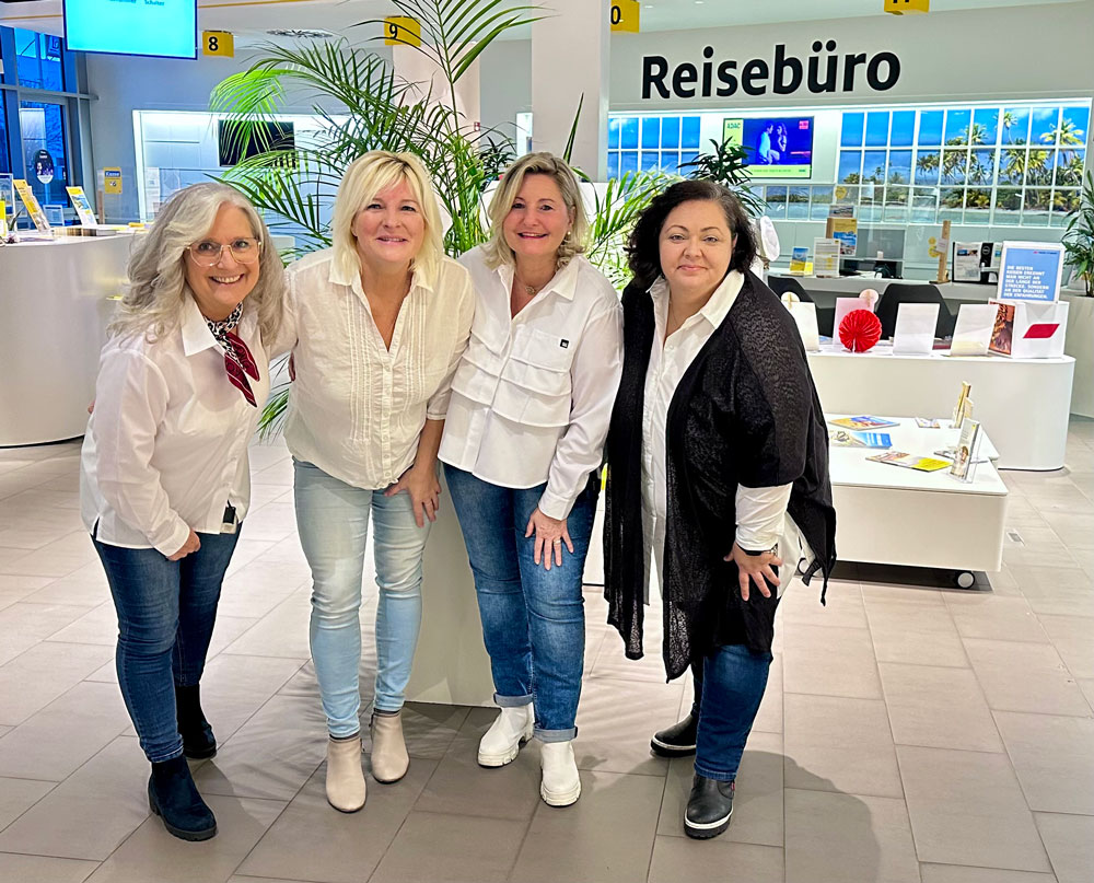 Vier freundlich lächelnde Frauen in weißer bzw. schwarzer Kleidung im Reisebüro
