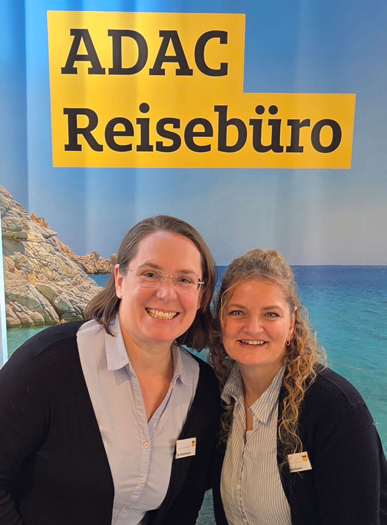 Zwei freundlich lächelnde Frauen vor einem ADAC Reisebüro Plakat mit Meeresmotiv