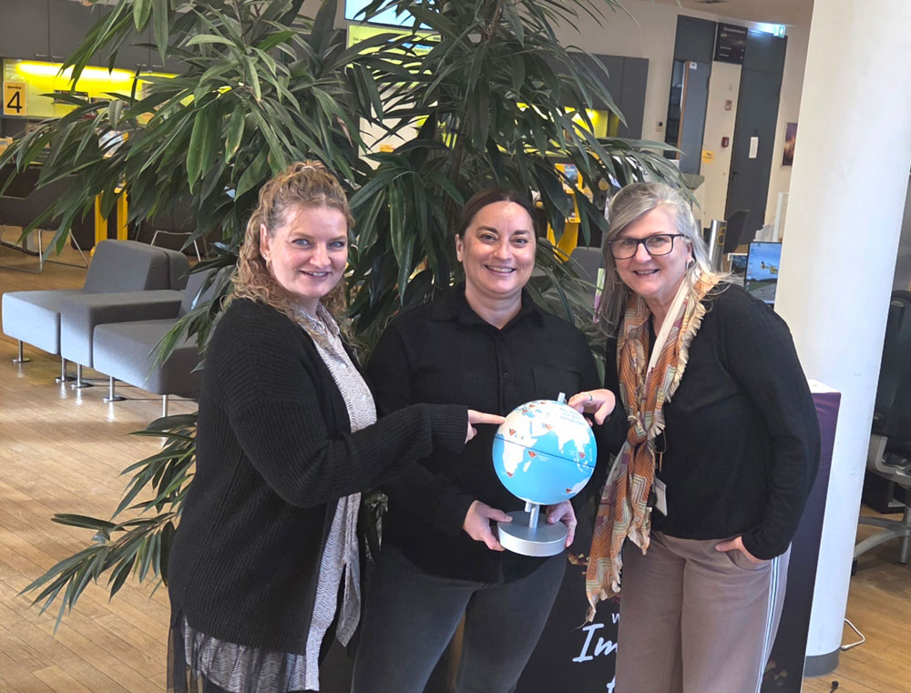 Drei lächelnde Frauen stehen neben einer Weltkugel in einem modernen Büro mit Pflanzen