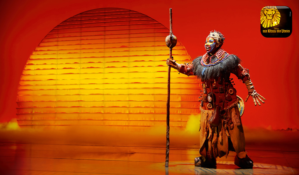 Eine Darstellerin im Löwenkostüm steht ausdrucksstark vor einer leuchtenden Sonne