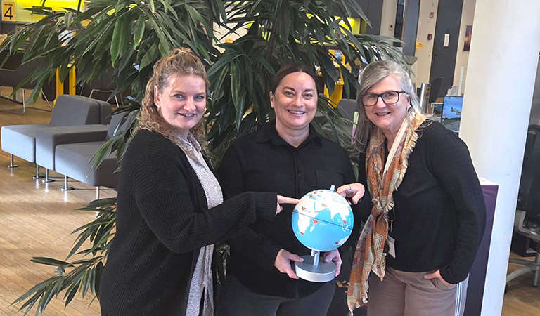 Drei lächelnde Frauen stehen vor einer großen Pflanze und halten gemeinsam einen Globus