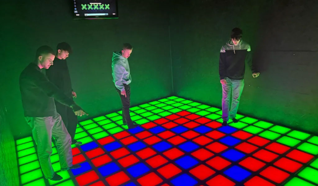 Vier junge Männer mit neutralem Gesichtsausdruck stehen auf einem leuchtenden LED-Feld mit grünen, roten und blauen Quadraten