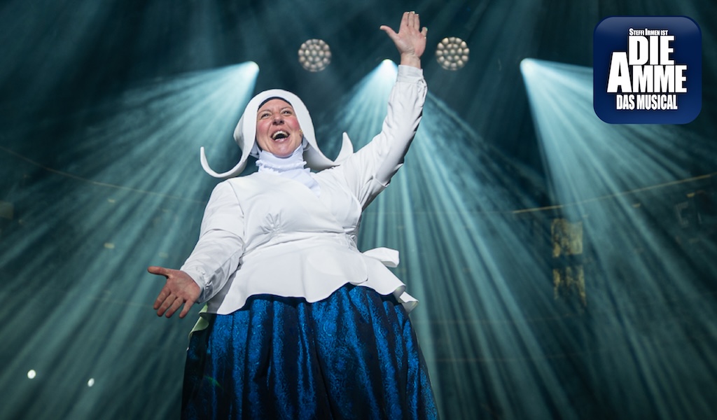 Eine Frau im weißen Haubenkostüm lacht herzlich auf einer dramatisch beleuchteten Bühne