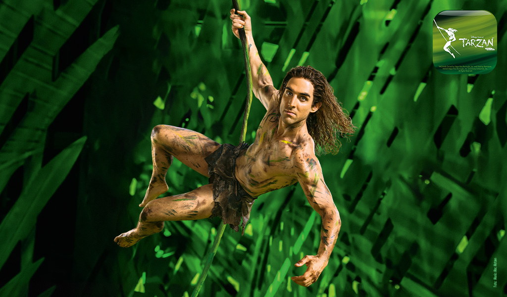 Ein muskulöser Mann mit langen braunen Haaren schwingt lächelnd an einer Liane vor grünem Dschungelhintergrund