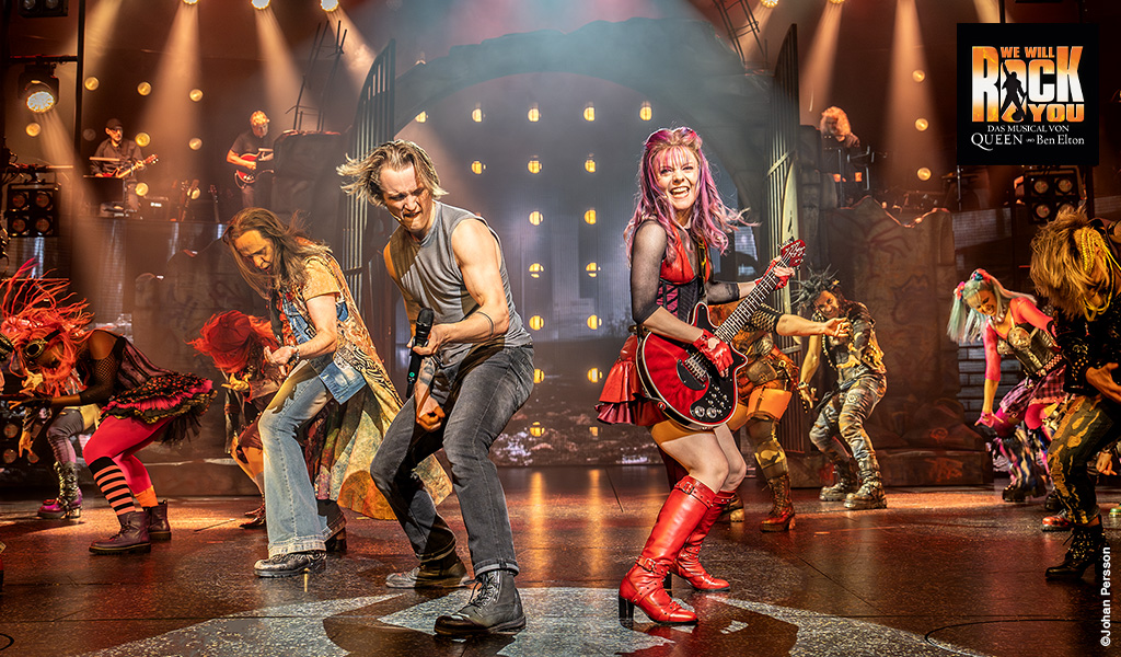 Eine Frau mit pinken Haaren und festlichem Make up spielt E Gitarre auf einer Bühne