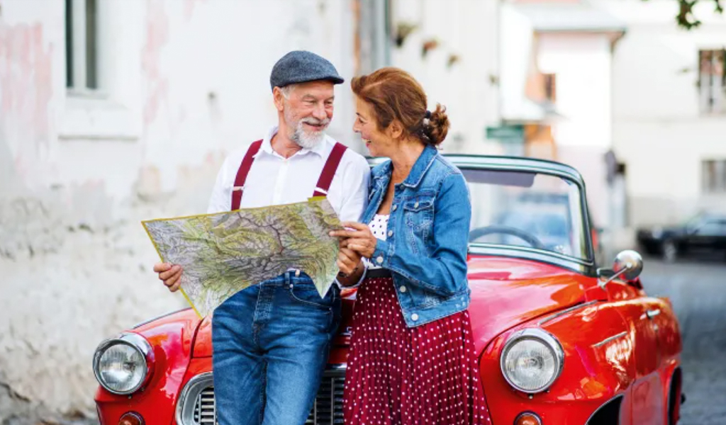 Ein älteres Paar lacht herzlich neben einem roten Oldtimer und hält eine große Landkarte
