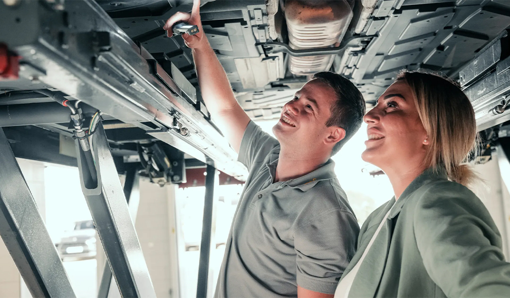 Zwei lächelnde Personen betrachten gemeinsam die Unterseite eines Fahrzeugs in einer Werkstatt