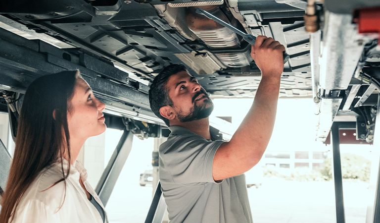 Ein Mann prüft konzentriert das Auto unter dem Wagen, eine Frau beobachtet aufmerksam