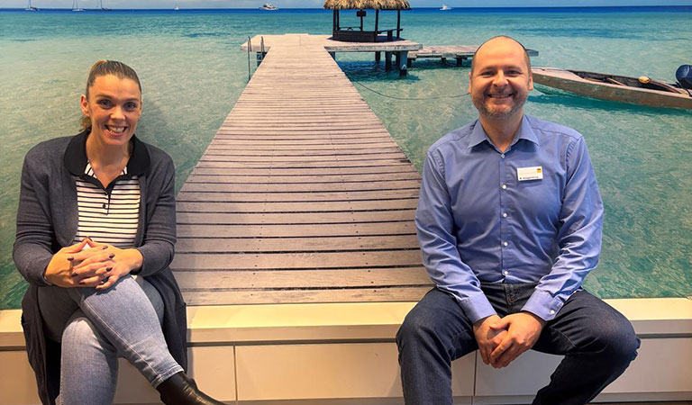 Zwei lächelnde Personen sitzen vor einem Foto eines Stegs am türkisblauen Meer