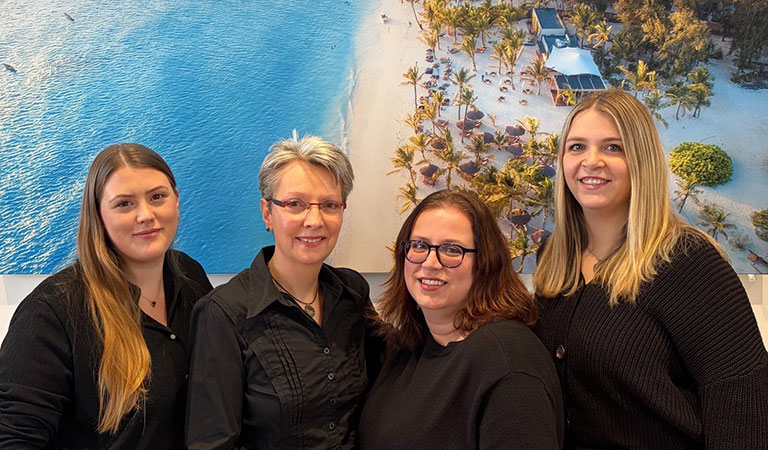 Vier Frauen mit freundlichen Gesichtern stehen vor einem Strandfoto in Blau und Beige