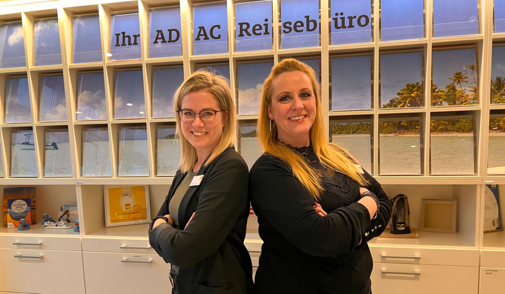 Zwei freundlich lachende Frauen mit Brille und blonden Haaren stehen Rücken an Rücken