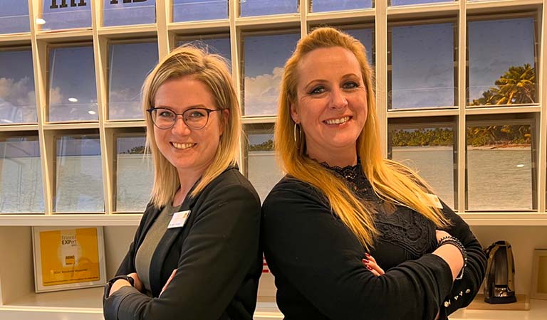 Zwei blonde Frauen stehen Rücken an Rücken und lächeln freundlich in einem Büro