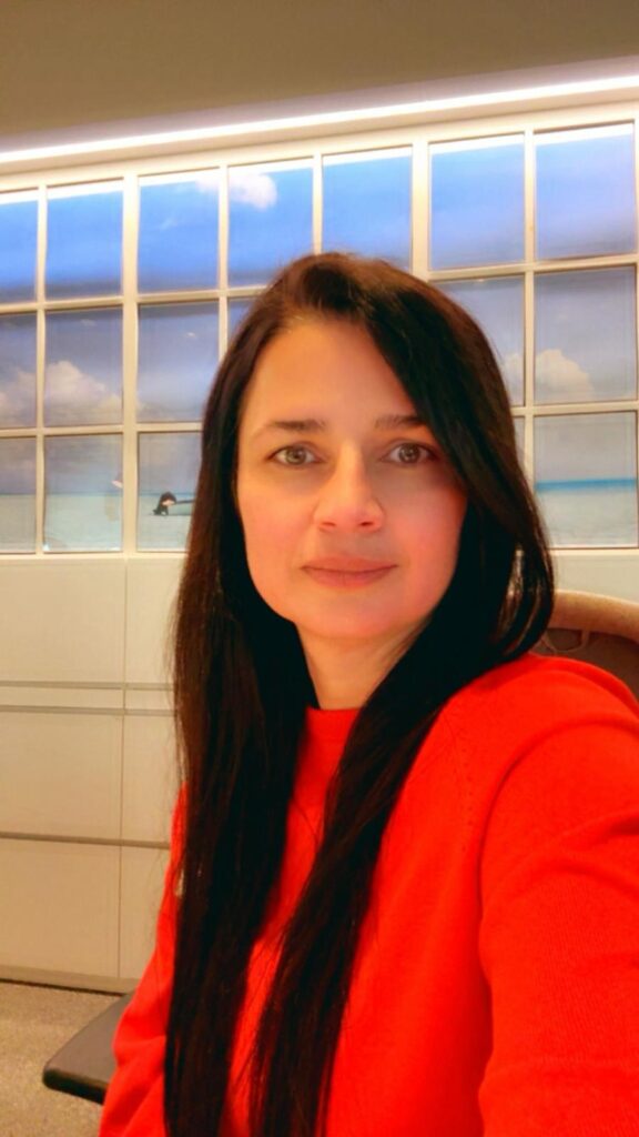 Eine Frau mit langen dunklen Haaren trägt einen leuchtend roten Pullover und sitzt vor einer Wand mit großen Fenstern und blauem Himmel