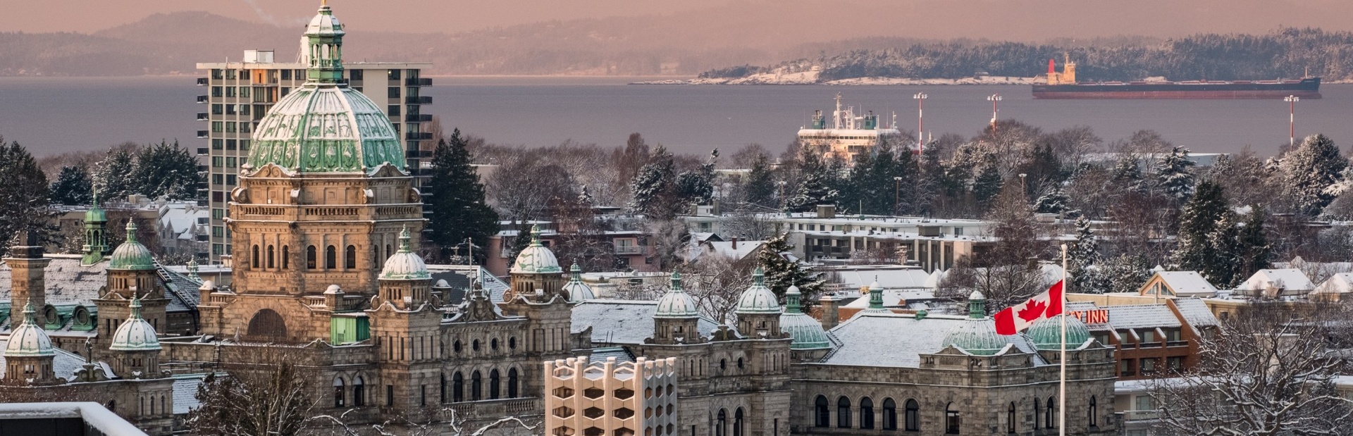 Das verschneite Parlamentsgebäude mit grünlicher Kuppel steht im Vordergrund, eine kanadische Flagge weht und im Hintergrund ist ein Schiff auf dem Wasser zu sehen