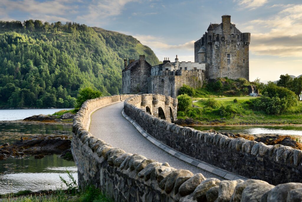 Eine alte Burg thront am Ende einer steinernen Brücke mit grün bewaldeten Hügeln