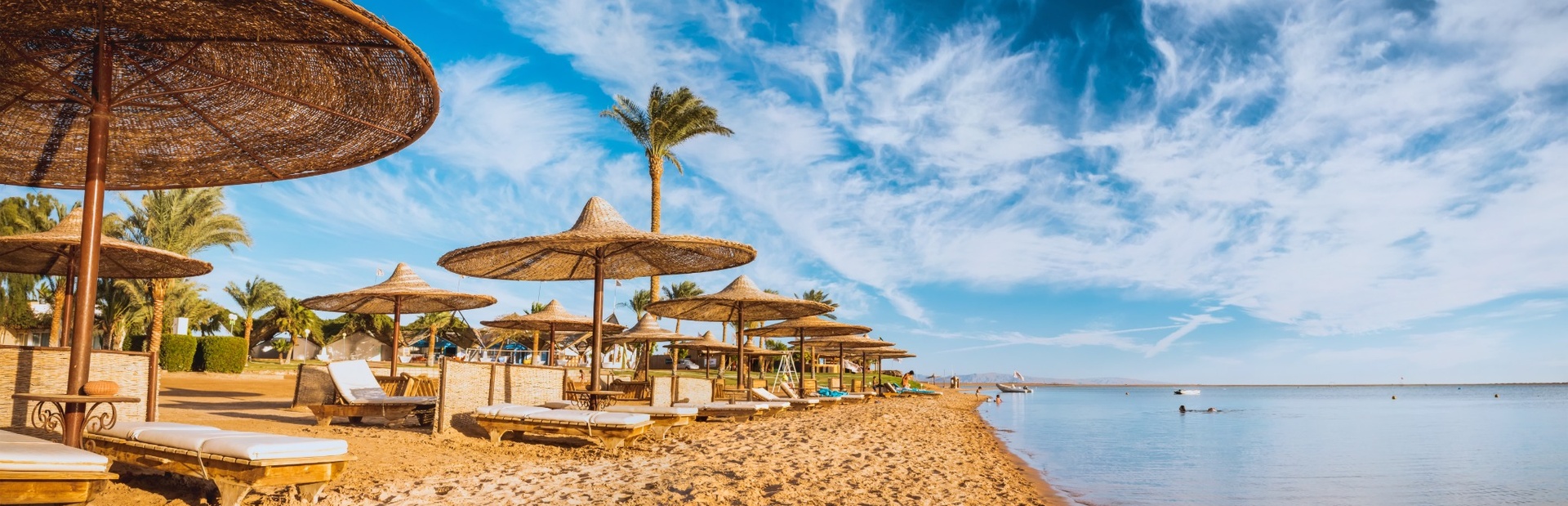 Breiter Sandstrand mit Sonnenliegen unter Strohdächern, klarem Meer und blauem Himmel