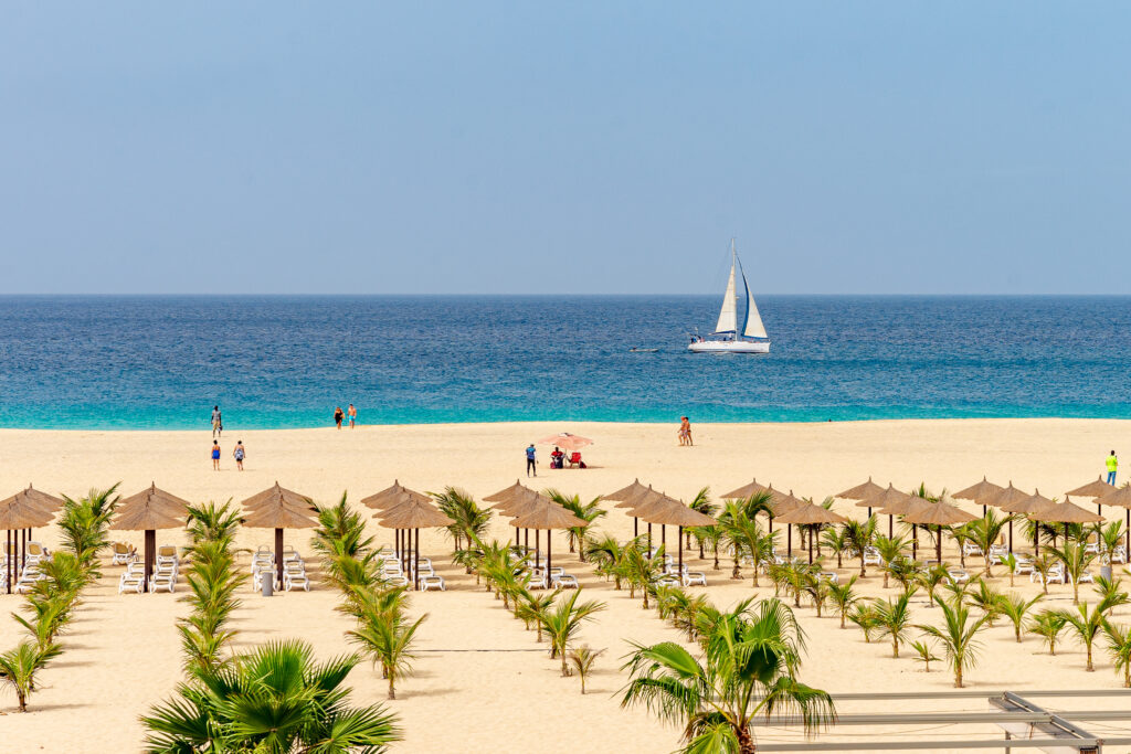 Ein sonniger Strand mit Palmen und Schirmen, Menschen spazieren, Segelboot im blauen Meer
