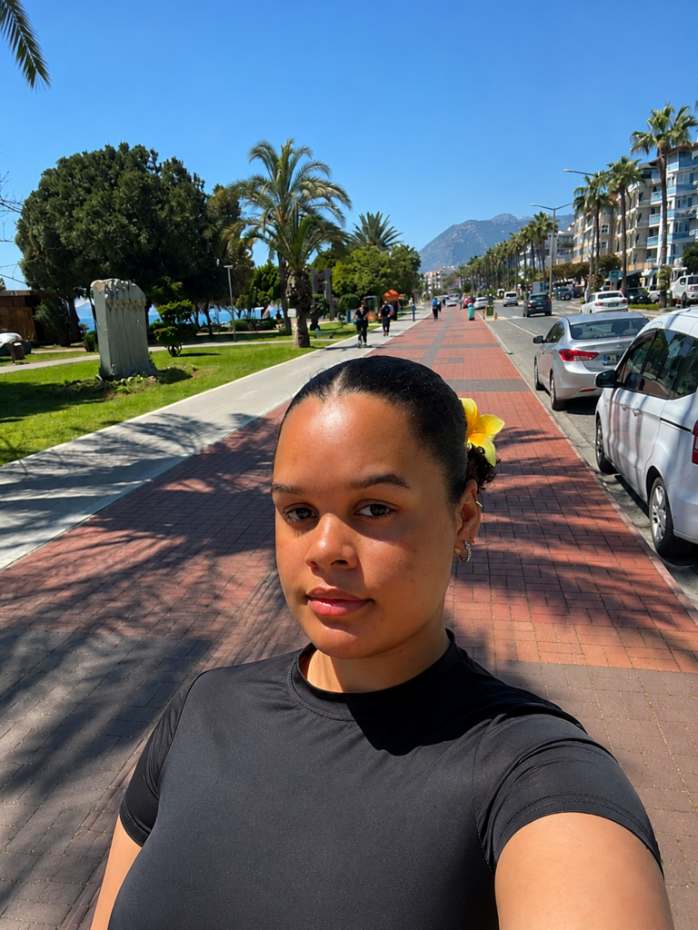 Eine Person mit dunklem Shirt und gelber Blume im Haar macht ein Selfie auf einer sonnigen Promenade mit Palmen und Bergen im Hintergrund