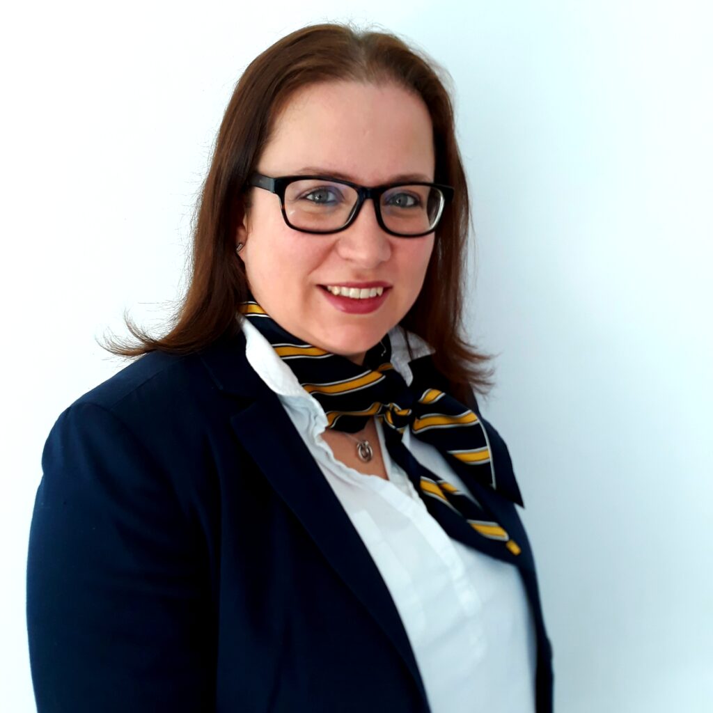 Eine Frau mit braunen Haaren trägt einen dunkelblauen Blazer und ein gestreiftes Halstuch