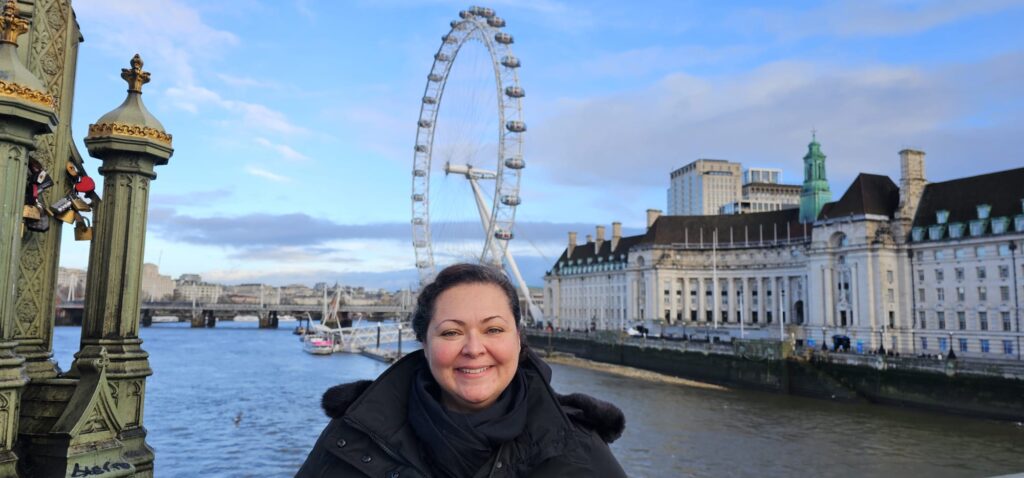 Mann mit dunkler Jacke steht lächelnd vor dem London Eye bei blauem Himmel