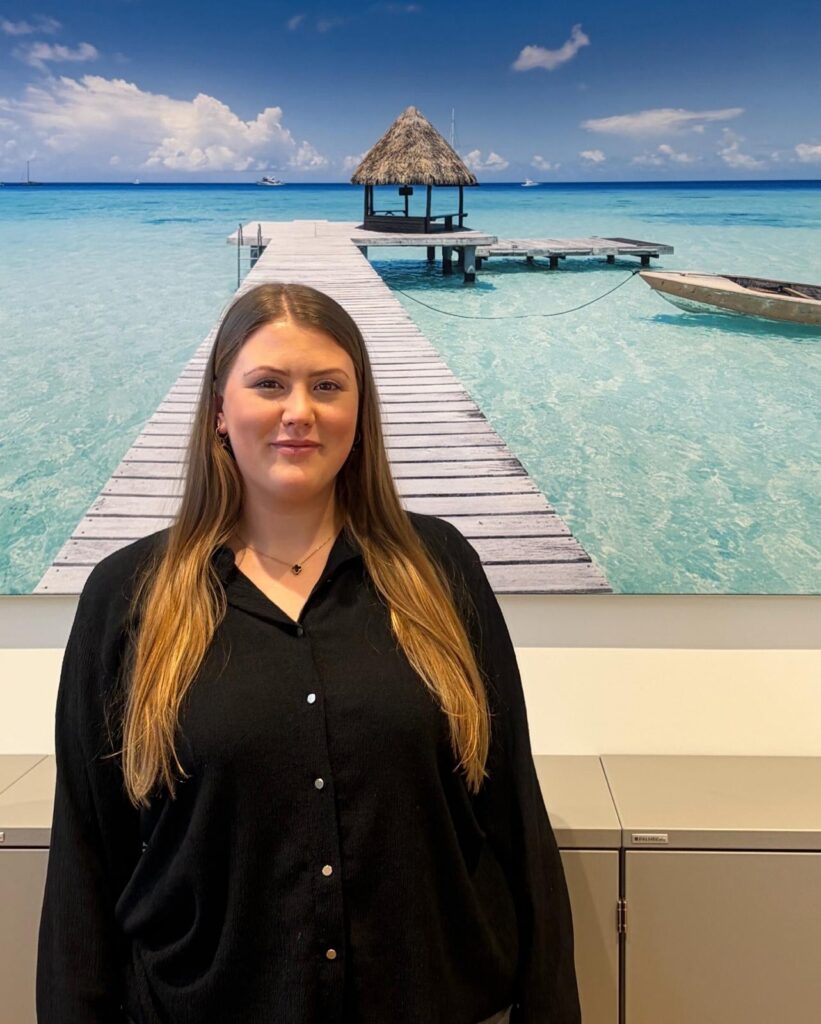 Eine Frau mit langem braunem Haar steht lächelnd vor einem tropischen Strandfoto