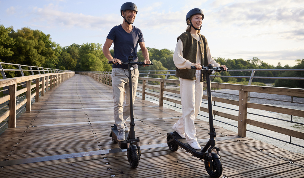 Zwei Personen mit neutralem Gesichtsausdruck fahren Elektroroller auf einer Holzbrücke