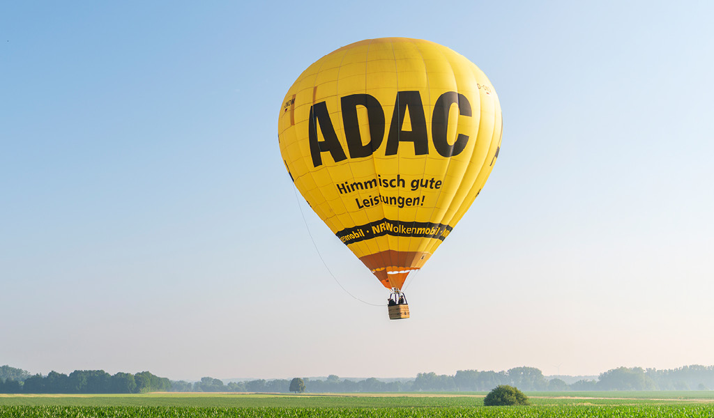 Ein leuchtend gelber ADAC Heißluftballon schwebt fröhlich über grüne Felder